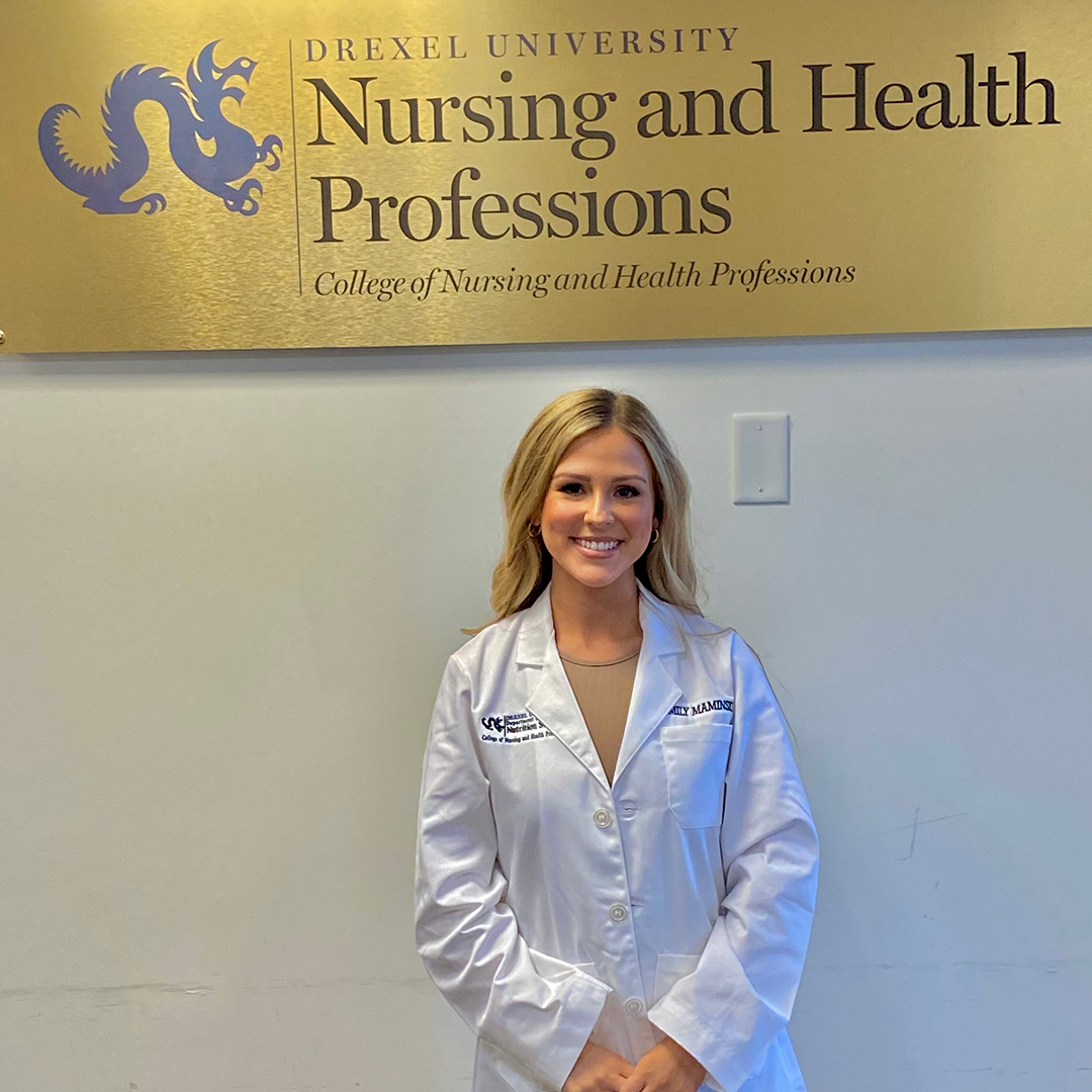 Emily Maminski stands in white coat in front of gold sign, 'Drexel University College of Nursing and Health Professions'
